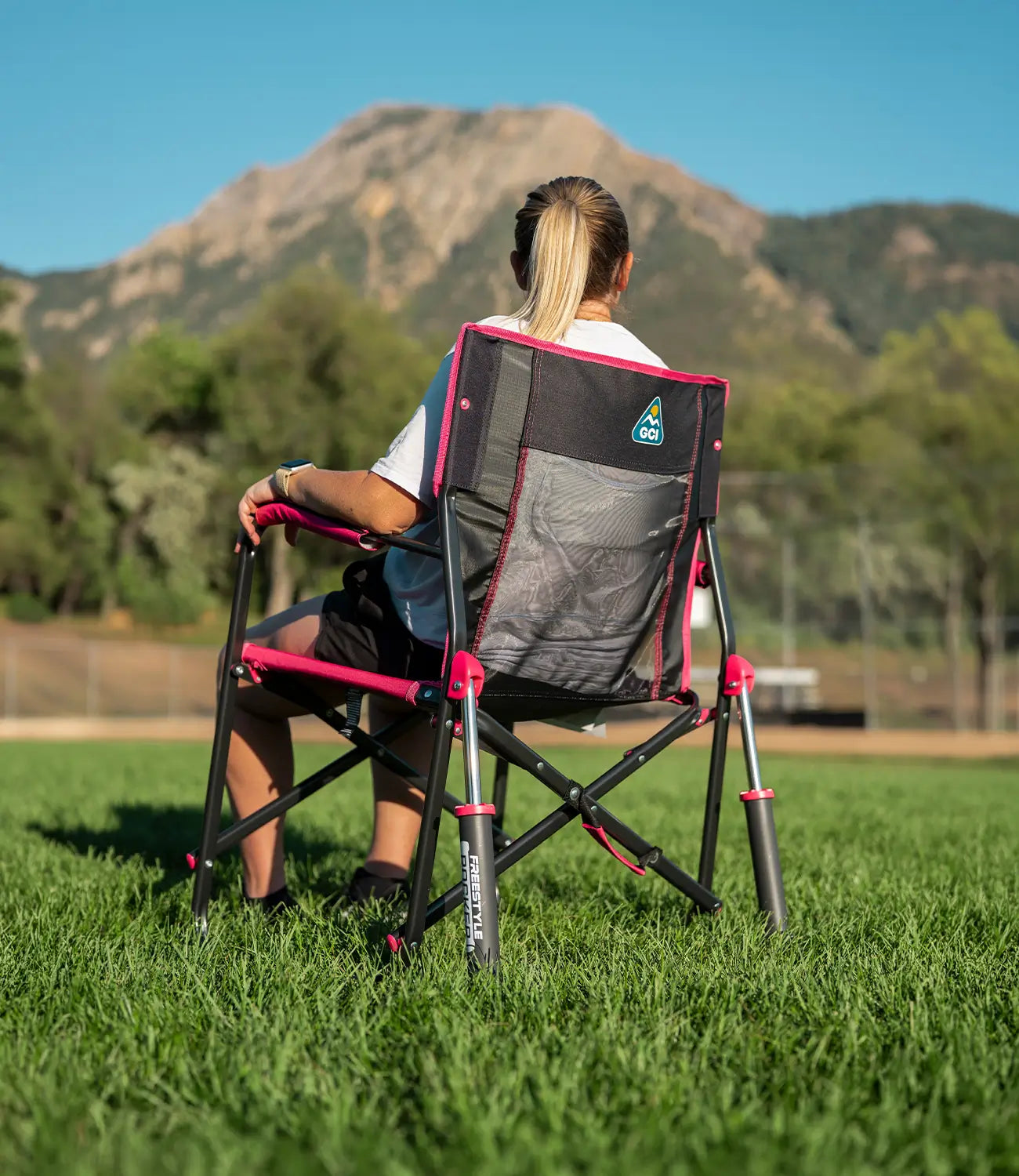 A woman sits in a Stowaway Rocker with a black and pink mesh back, viewed from behind as she looks toward a mountain in a grassy field.