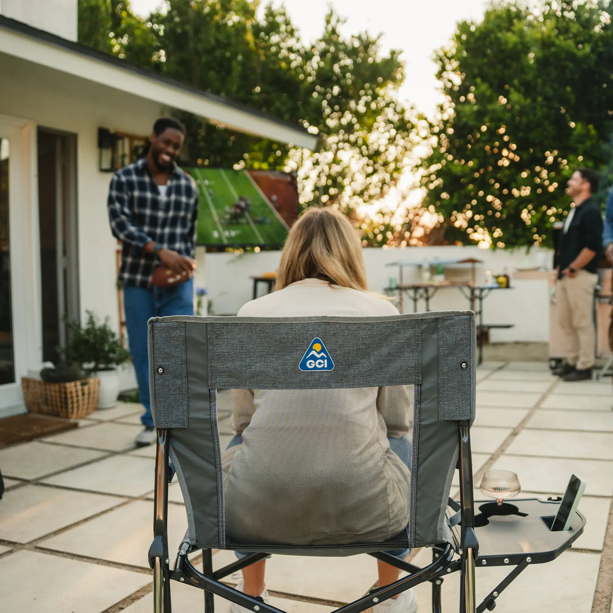 A woman sitting in a gray freestyle rocker with side table watching a football game on television. 