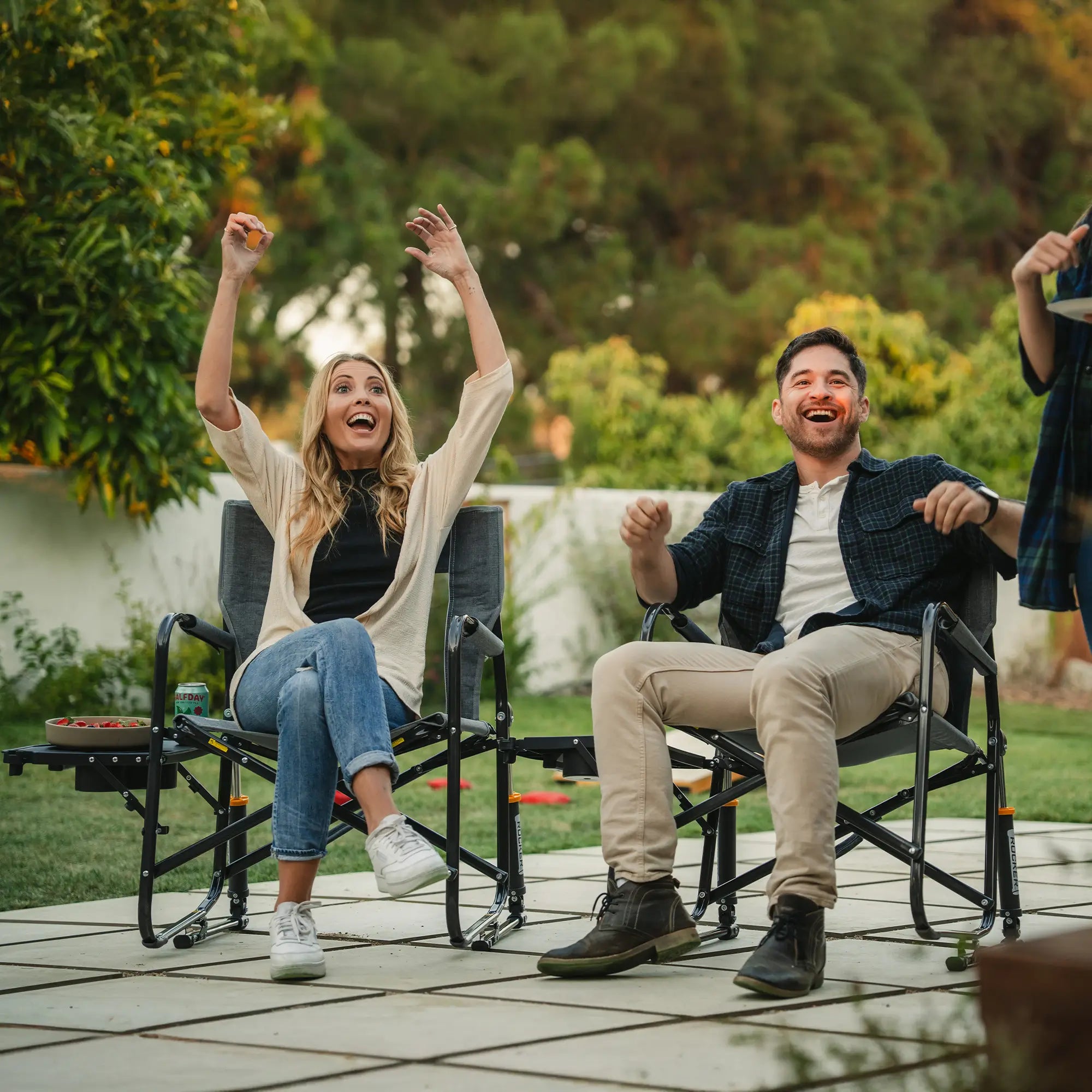 Two people cheering and smiling as they sit in their freestyle rocker with side table on a patio. 