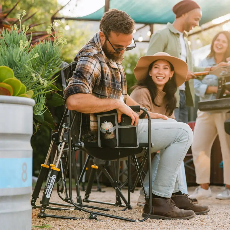 Man seated in a black Freestyle Rocker XL at a gathering, using the built-in gear pockets while others socialize nearby.