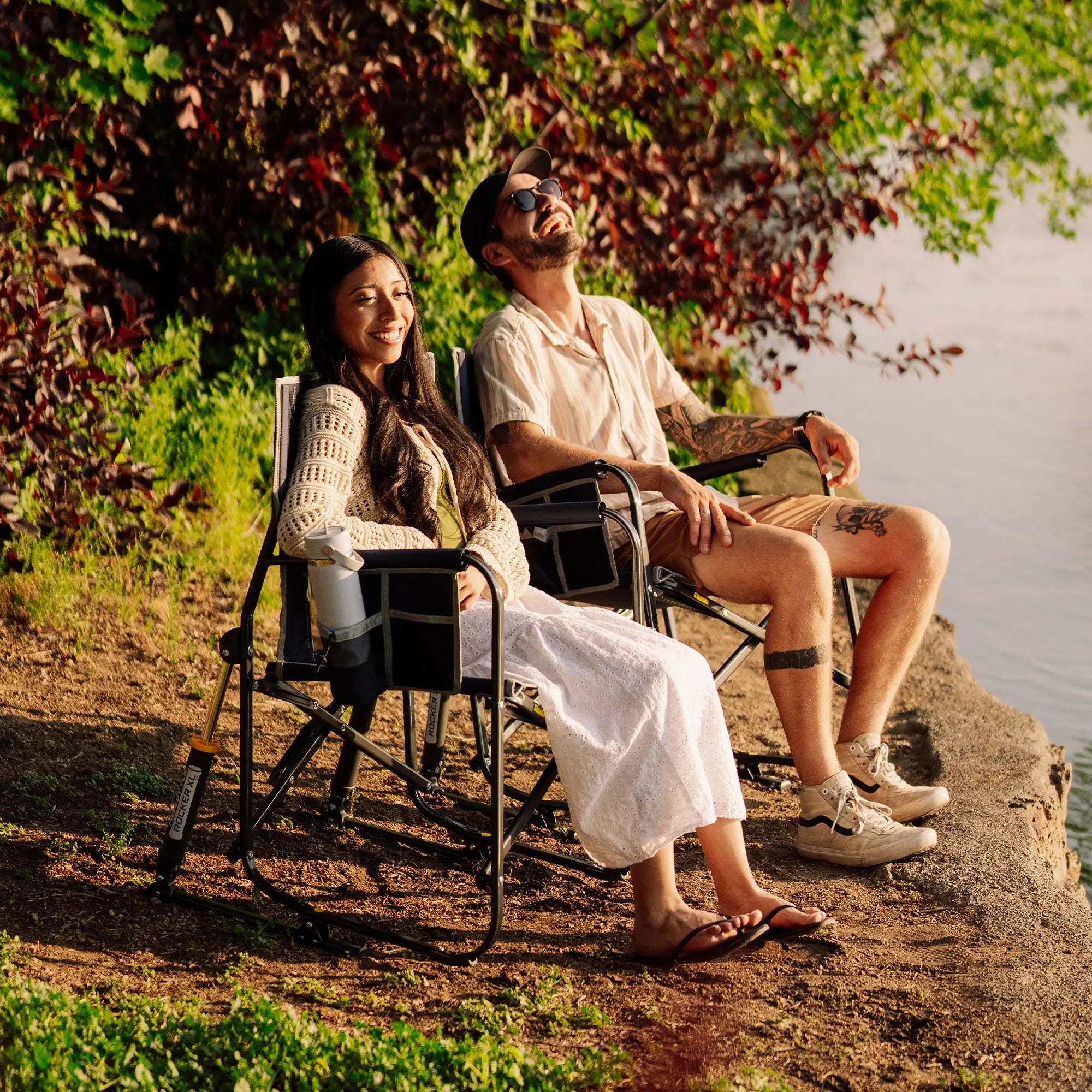 A man and a woman relax in black freestyle rocker xl chairs on a rocky lakeside, surrounded by lush green foliage and warm sunlight.