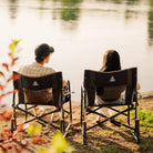 Two people sitting in black freestyle rocker xl chairs facing a lake, photographed from behind as they relax outdoors by the water.