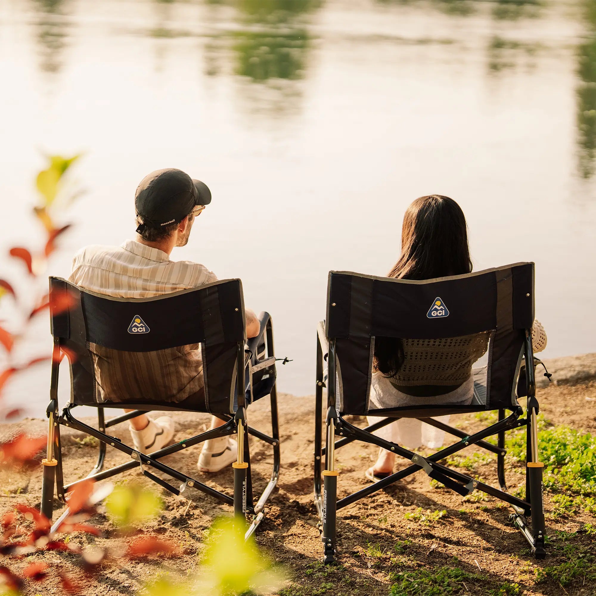 Two people sitting in black freestyle rocker xl chairs facing a lake, photographed from behind as they relax outdoors by the water.