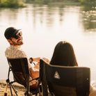 Two people sitting in black freestyle rocker xl chairs by a lake at sunset, viewed from behind the seated woman.
