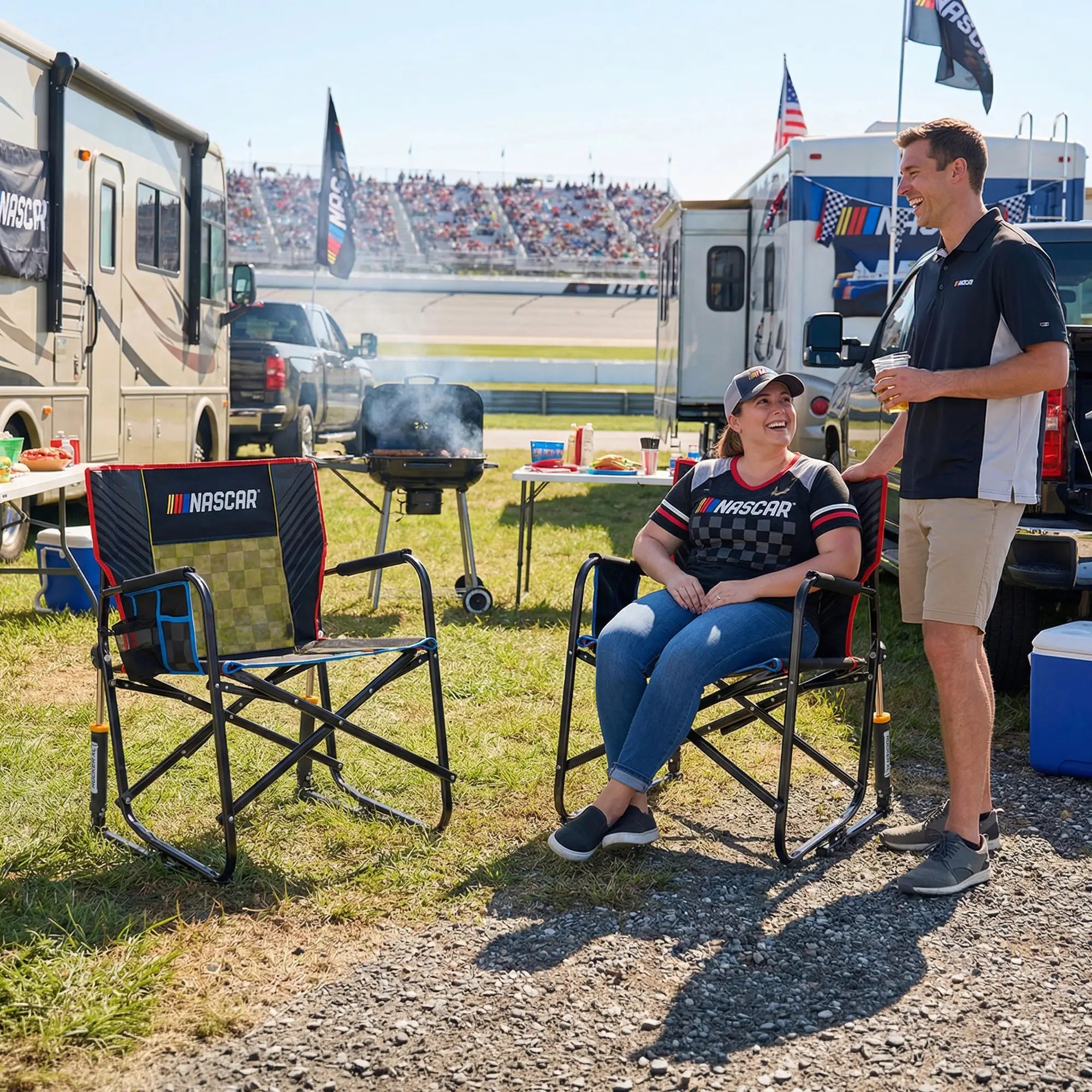 A man and a woman relax in black and red NASCAR Freestyle Rocker XL chairs at an outdoor racetrack event, with RVs, a grill, and a grandstand in the background.