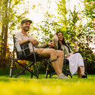 Two people sitting in Freestyle Rocker XL chairs on a grassy lawn outdoors, with trees and bright sunlight in the background.