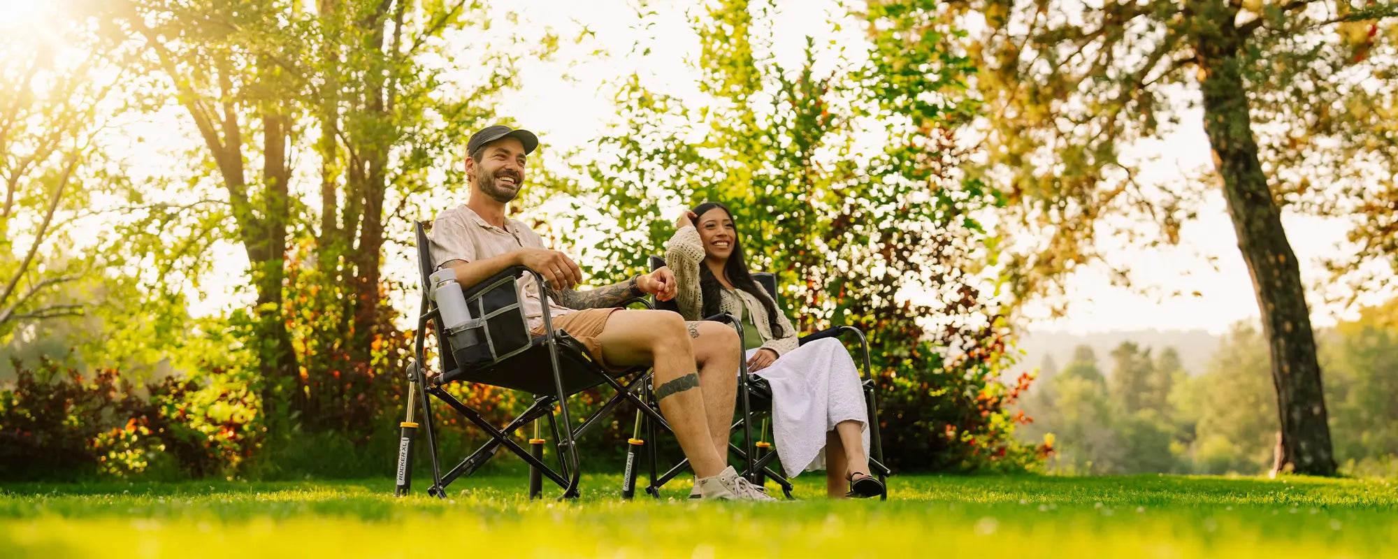 A man and a woman relax in black freestyle rocker xl chairs on a grassy lawn in a sunny park, surrounded by lush green trees and foliage.
