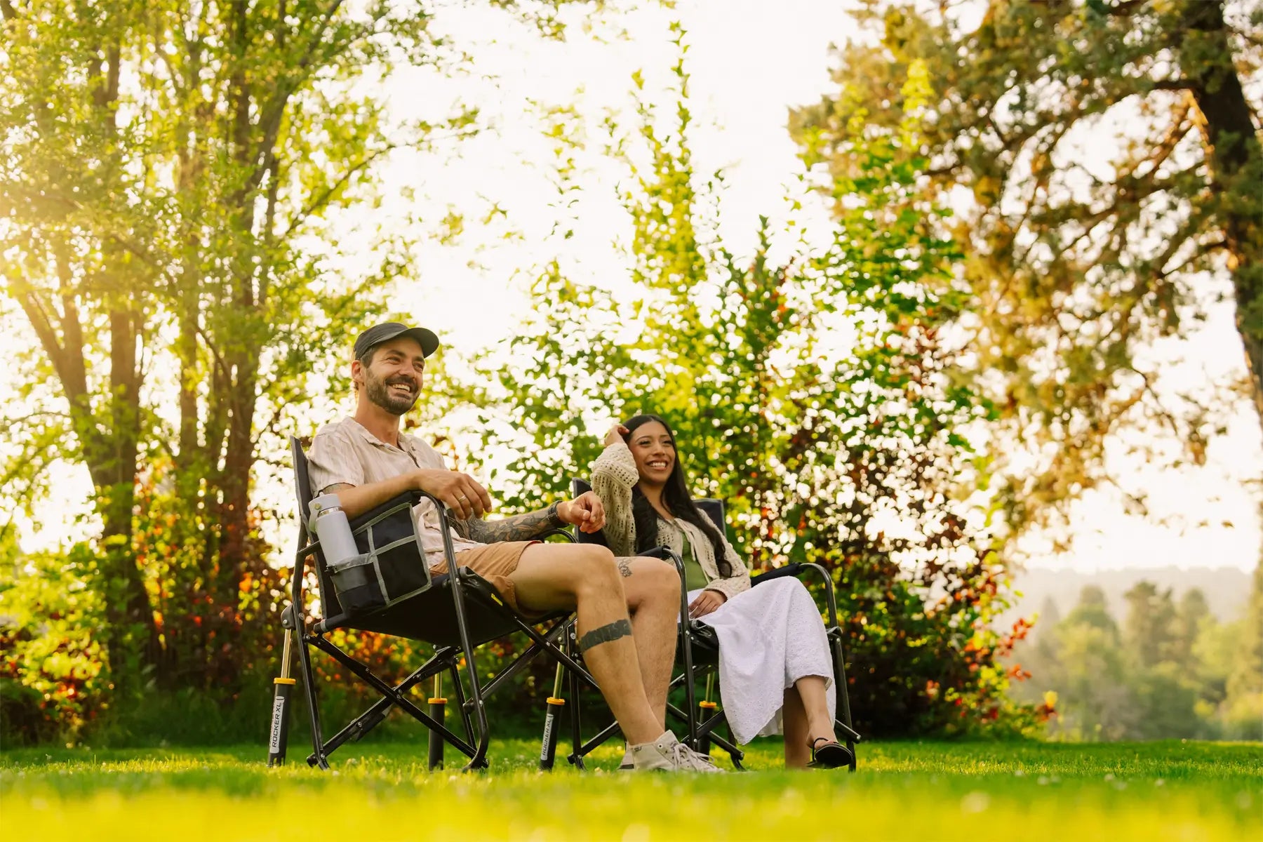 A man and a woman relax in black freestyle rocker xl chairs on a grassy lawn in a sunny park, surrounded by lush green trees and foliage.