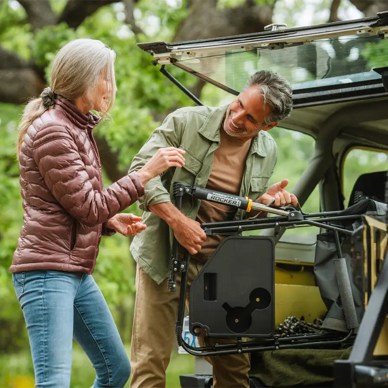 Man unloading a folded Freestyle Rocker with Side Table in heathered pewter from a vehicle while smiling with a woman nearby.