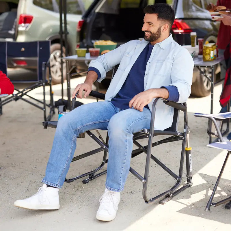 Man seated and smiling in a black Freestyle Rocker XL with Side Table at a tailgate, surrounded by cars and food tables.