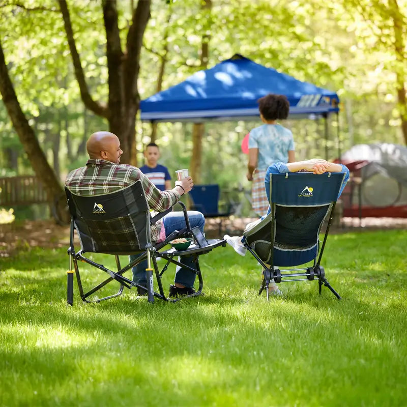 Two people relaxing in Freestyle Rocker XL chairs at a wooded campsite, with kids playing nearby under a blue canopy tent.