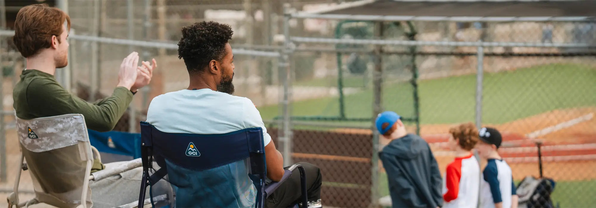 Two men sit in GCI Outdoor Freestyle Rocker Elite chairs at a baseball field.