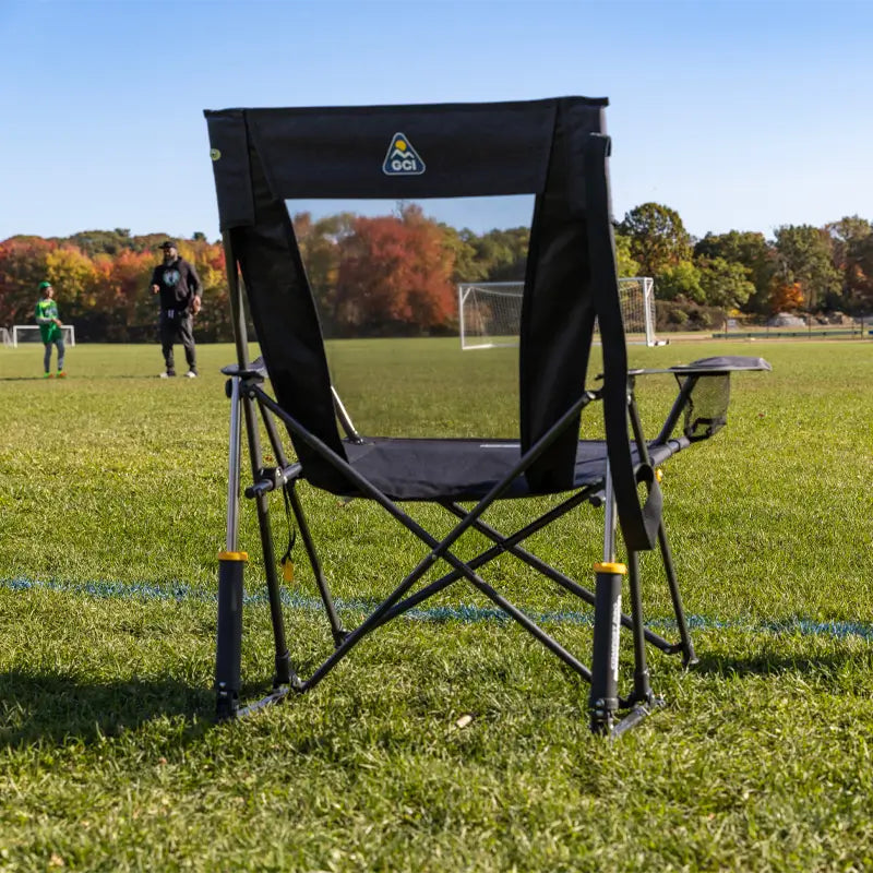 A Comfort Pro Rocker XL sits on the sidelines of a soccer field, offering a comfortable spot for spectators. The chair features a high backrest, padded armrests, and a breathable mesh panel for support during long outdoor events.