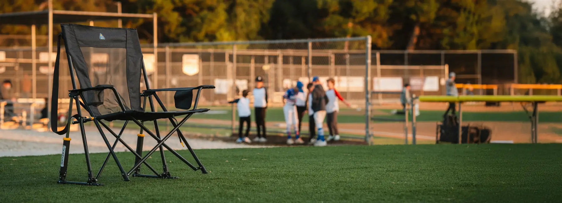 A Comfort Pro Rocker XL chair sits on the edge of a baseball field, with a game happening in the background. This oversized rocking chair features a breathable mesh back, sturdy frame, and smooth rocking motion, perfect for outdoor sports and events.