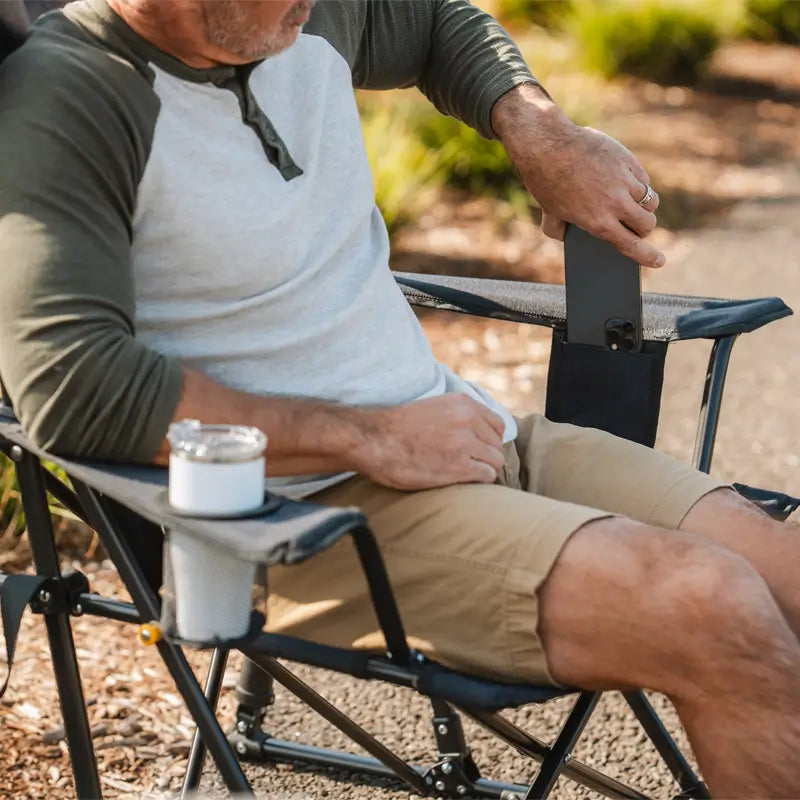 A man sits in a Comfort Pro Rocker XL, placing his phone into a built-in side pocket while his drink rests in the cup holder. The chair's thoughtful design provides convenient storage for essentials, making outdoor relaxation even easier.