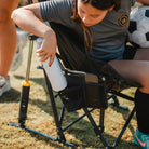 A young soccer player sits in a GCI Outdoor Pod Rocker Elite, a portable rocking camp chair, placing a water bottle into its mesh pocket.