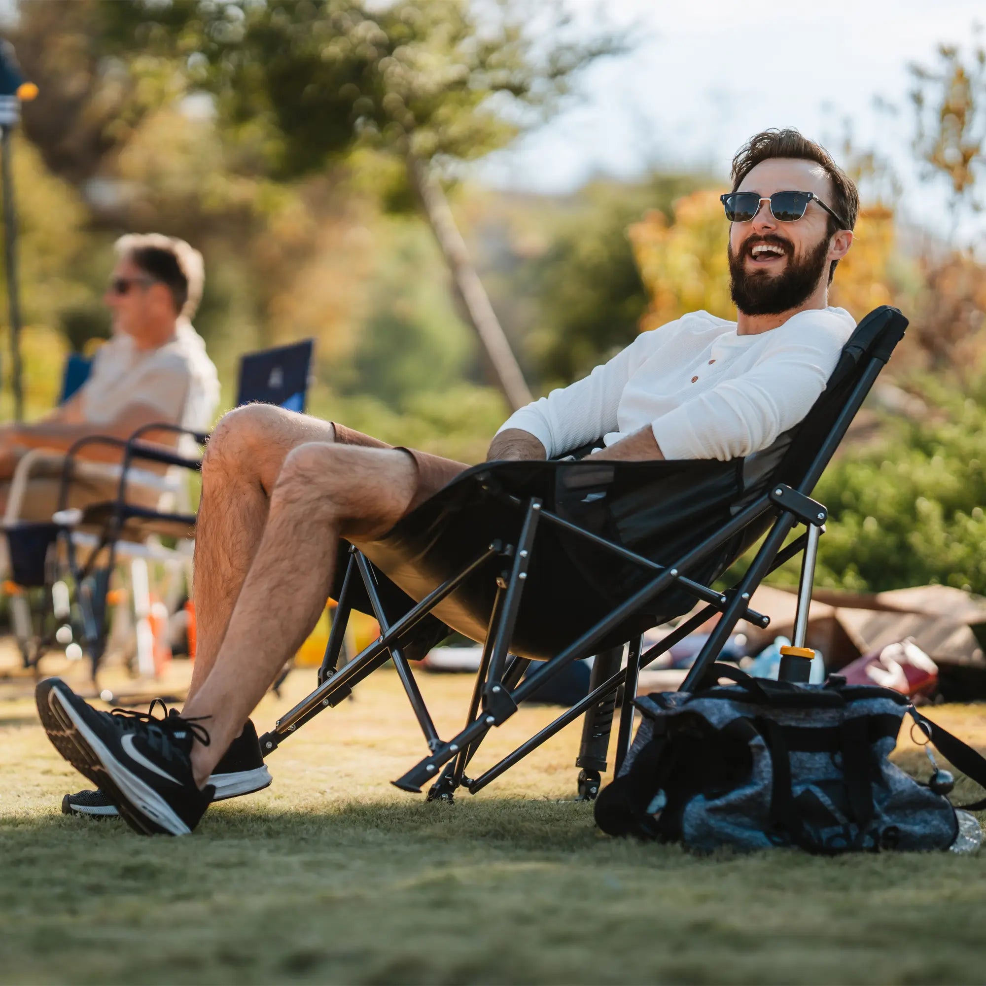 A man in sunglasses relaxes in a GCI Outdoor Pod Rocker Elite, a portable rocking camp chair, smiling while enjoying an outdoor event.