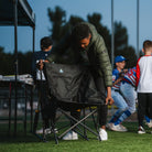 A man unfolds a GCI Outdoor Pod Rocker Elite at a nighttime baseball game, setting up the rocking camp chair on the field.