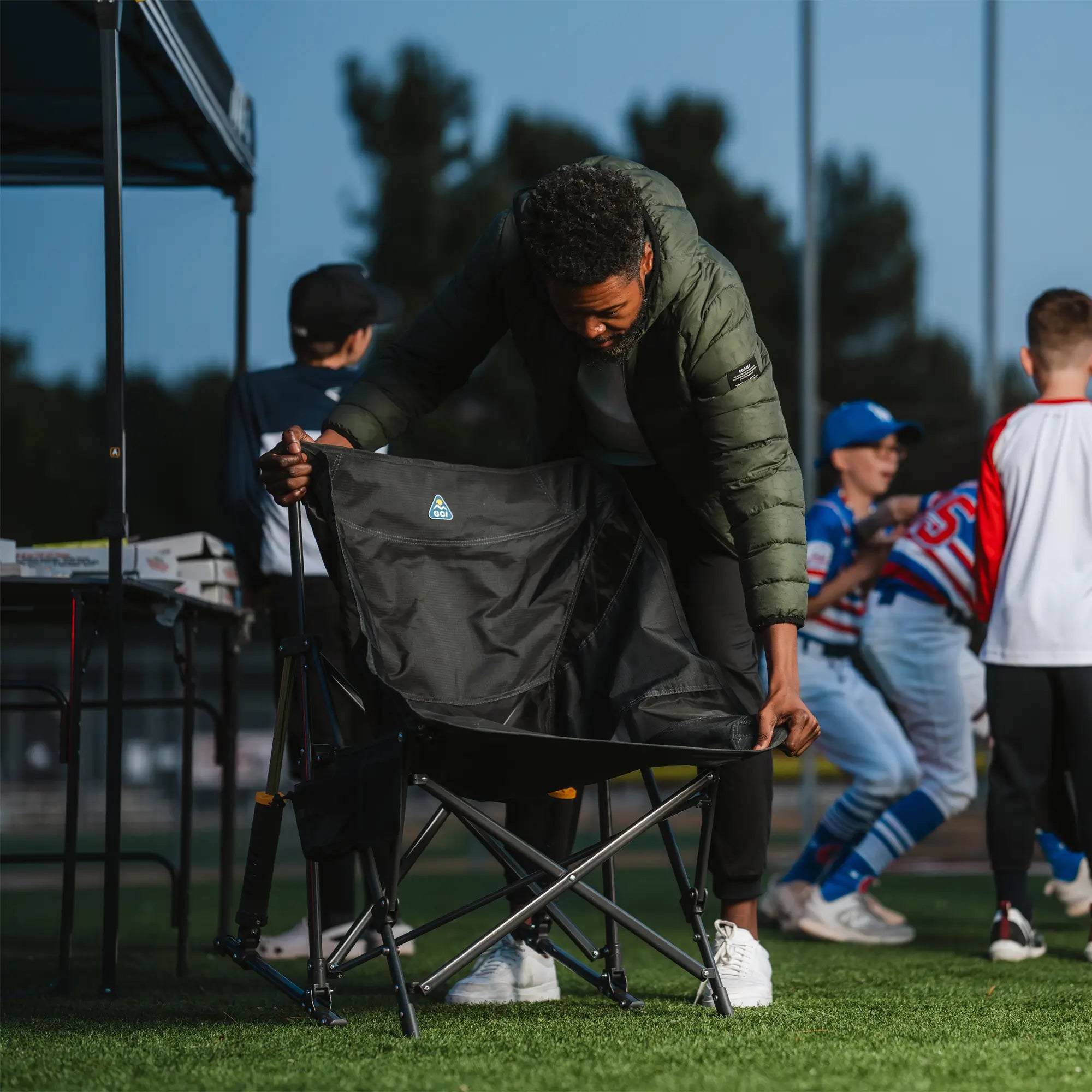 A man unfolds a GCI Outdoor Pod Rocker Elite at a nighttime baseball game, setting up the rocking camp chair on the field.