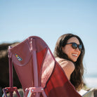 A smiling woman in sunglasses sits in a GCI Outdoor Pod Rocker Elite, enjoying a sunny beach day with the ocean in the background.