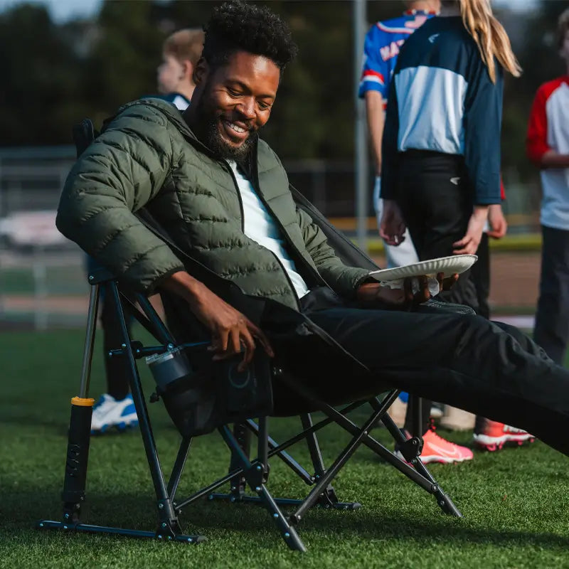 A man reclines in a GCI Outdoor Pod Rocker Elite, smiling as he enjoys a meal at a nighttime sports event.