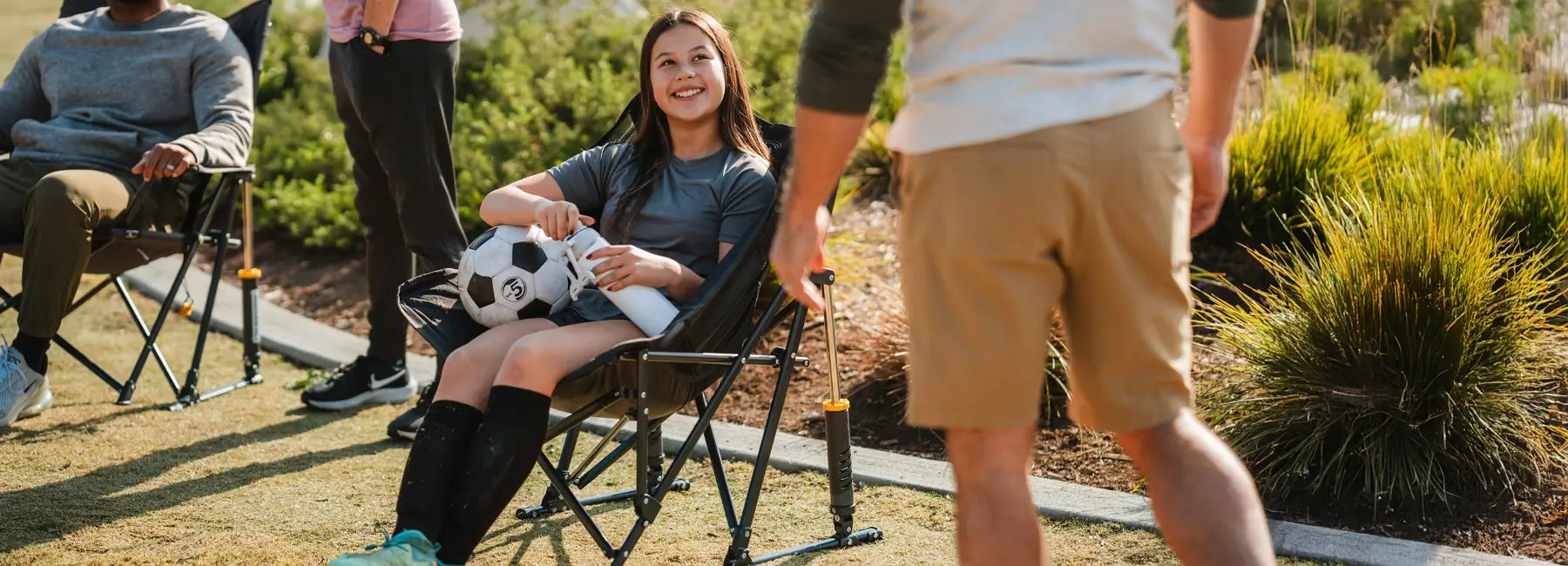 A young soccer player sits in a GCI Outdoor Pod Rocker Elite, holding a soccer ball and water bottle while smiling at someone standing nearby.