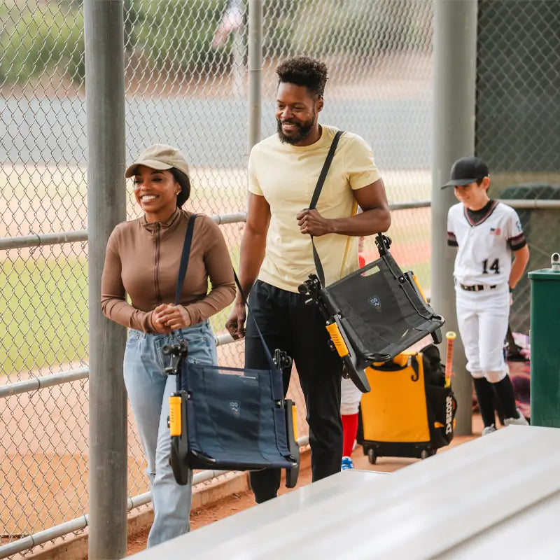 Couple carrying GCI Outdoor Stadium Rock-Cliner chairs by shoulder straps, heading to a baseball bleacher with portable seats.