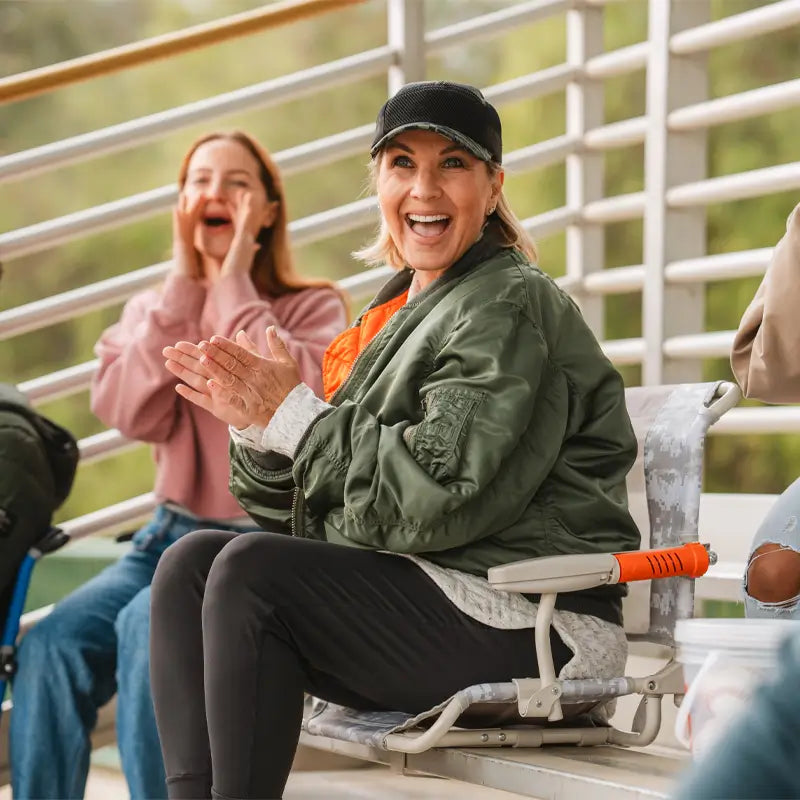 A woman cheering on a bleacher in her GCI Outdoor Stadium Rock-Cliner with orange armrests and a foldable, rocking design.