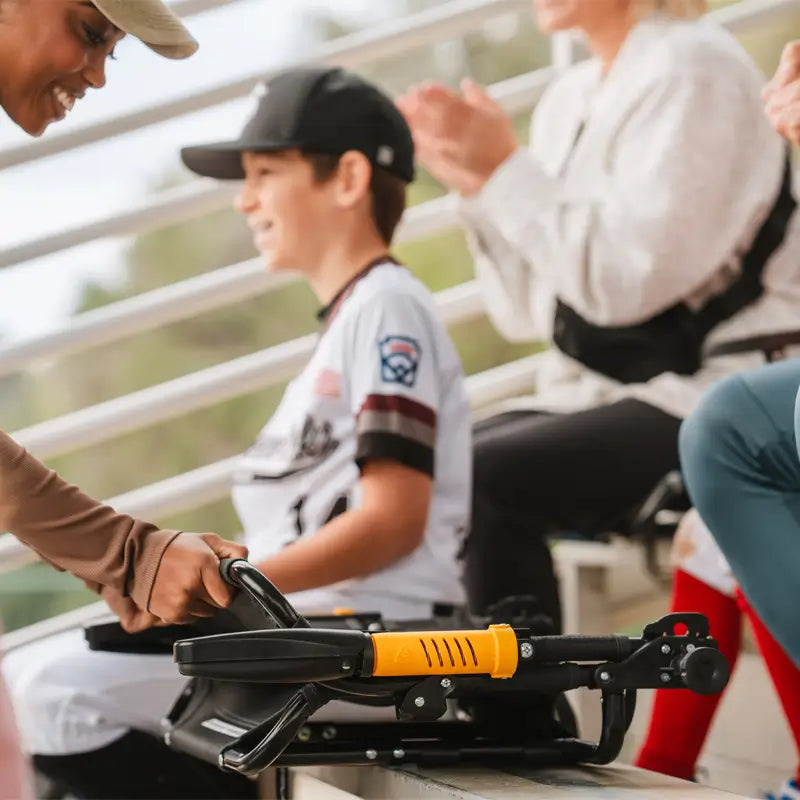 GCI Outdoor Stadium Rock-Cliner, a foldable rocking stadium seat with gold armrests, on a bleacher at a youth baseball game.