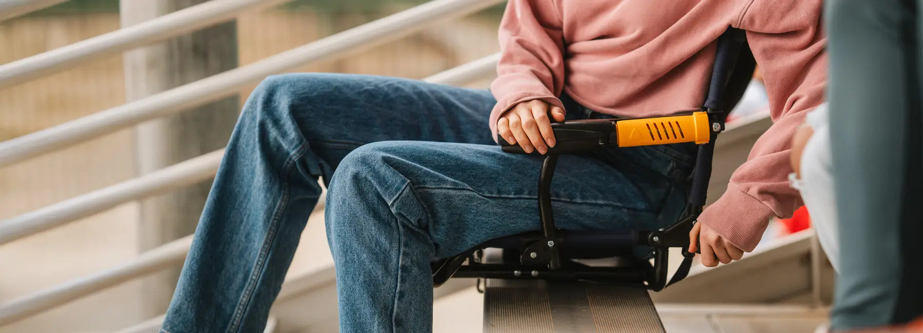 A person in pink adjusts a GCI Outdoor Stadium Rock-Cliner on bleachers, highlighting its orange armrest and foldable design.