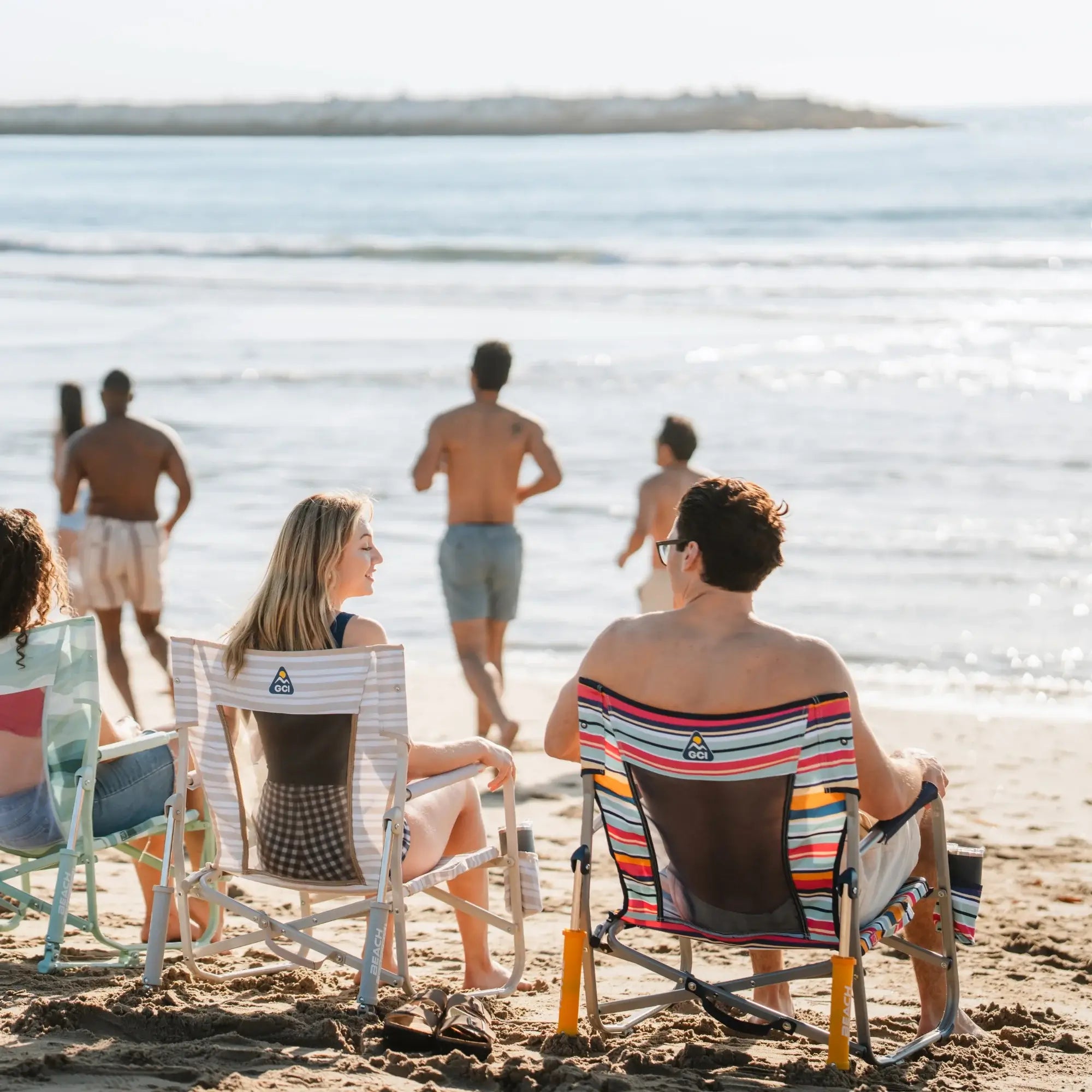 A group sitting at the beach in their colorful beach rockers while people run in front of them.