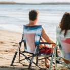 A man sitting in a blue beach rocker near the ocean.