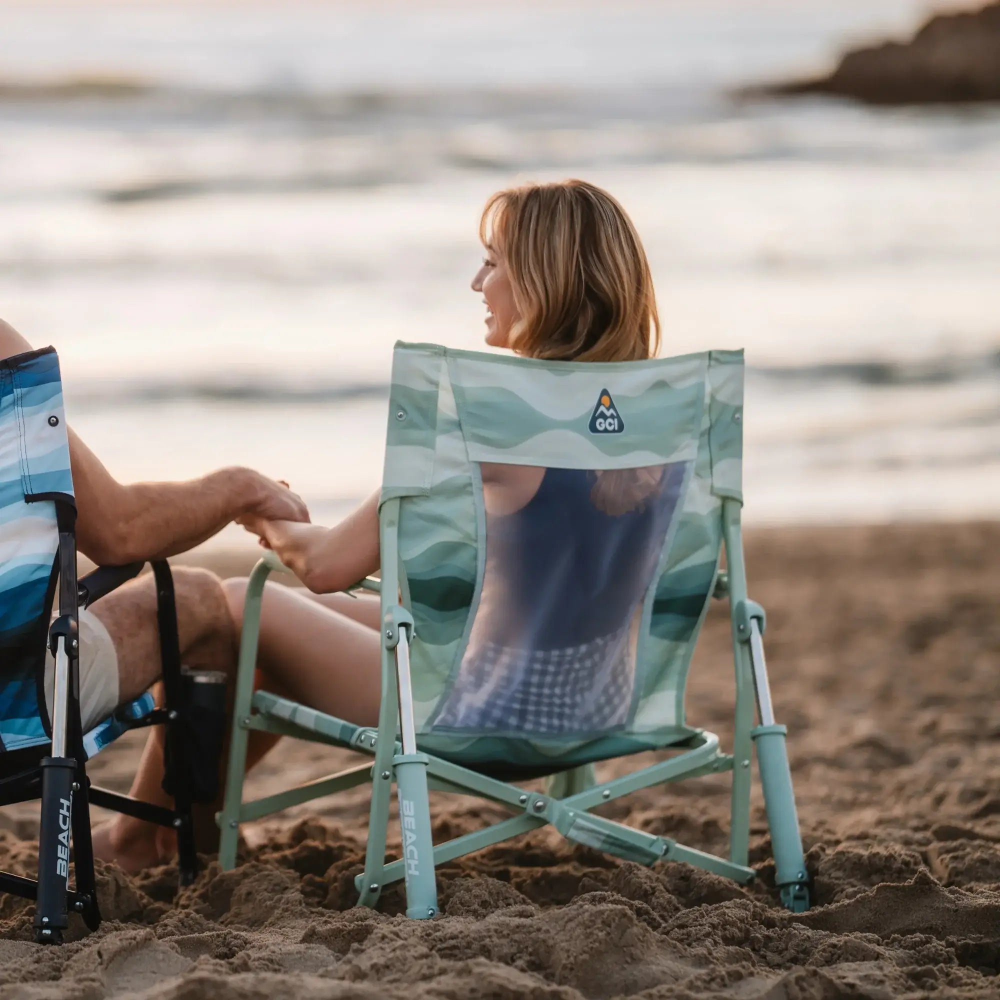 A woman sitting in a teal beach rocker while talking to a man on the beach.