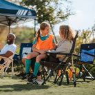 Two young girls sit together in a Comfort Pro Rocker XL at a soccer game, smiling and chatting. The sturdy, oversized rocking chair provides a comfortable spot for spectators, with padded armrests and a supportive mesh back.