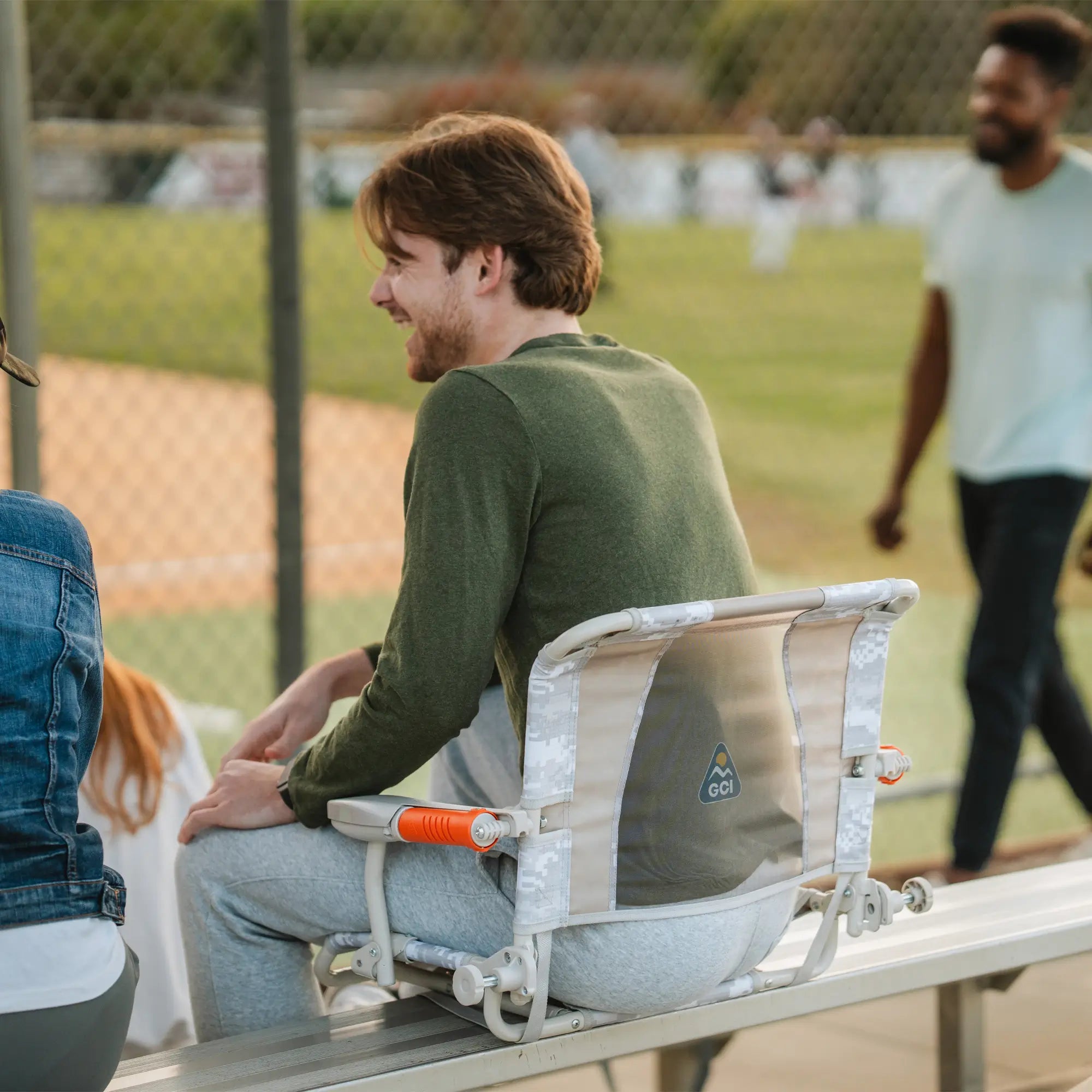 A man sits comfortably on a camo GCI Outdoor Stadium Rock-Cliner with orange armrests at a baseball game.