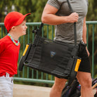 Man carrying a black GCI Outdoor Stadium Rock-Cliner with gold armrests by a shoulder strap, showing its portable design.