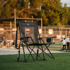 A Comfort Pro Rocker XL chair sits on a grassy field near a baseball game, bathed in warm evening light. The sturdy, oversized rocker features a breathable mesh back, padded armrests, and a smooth rocking mechanism for ultimate outdoor comfort.