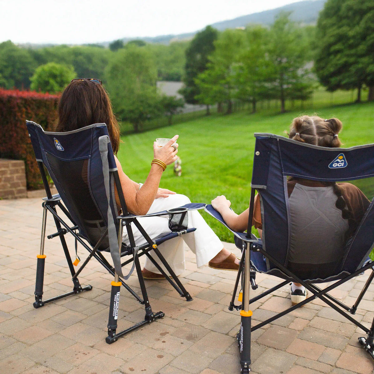 Two people sitting in camping chairs on a patio with a scenic view of trees and grass.