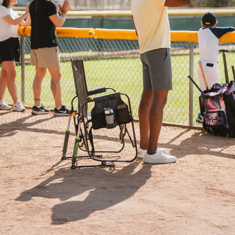 Man standing cheering on baseball players with a stowaway rocker next to him.