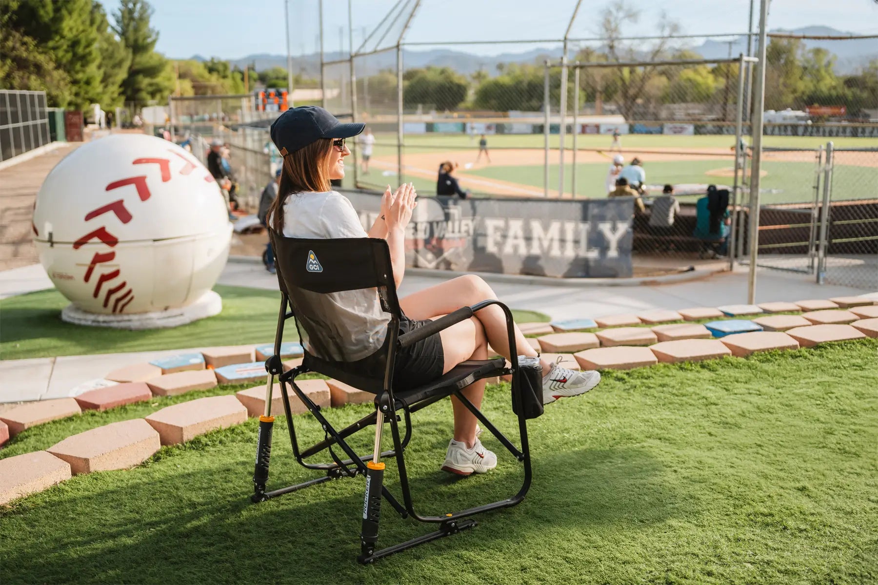 Woman sitting in freestyle rocker elite cheering on baseball team while positioned next to a baseball sculpture.