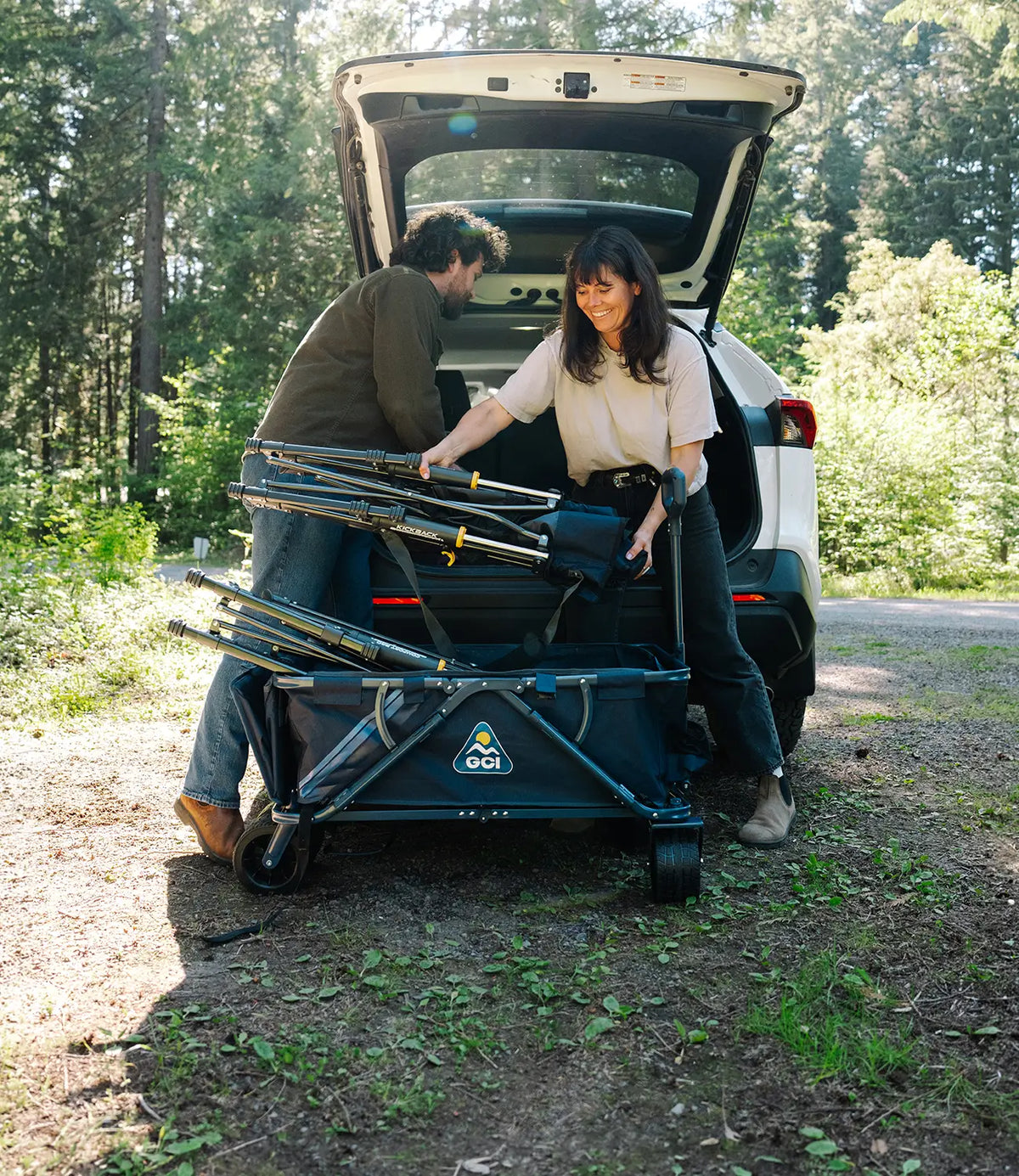 A man and a woman loading camping equipment into the open trunk of a white SUV on a dirt road surrounded by trees. 