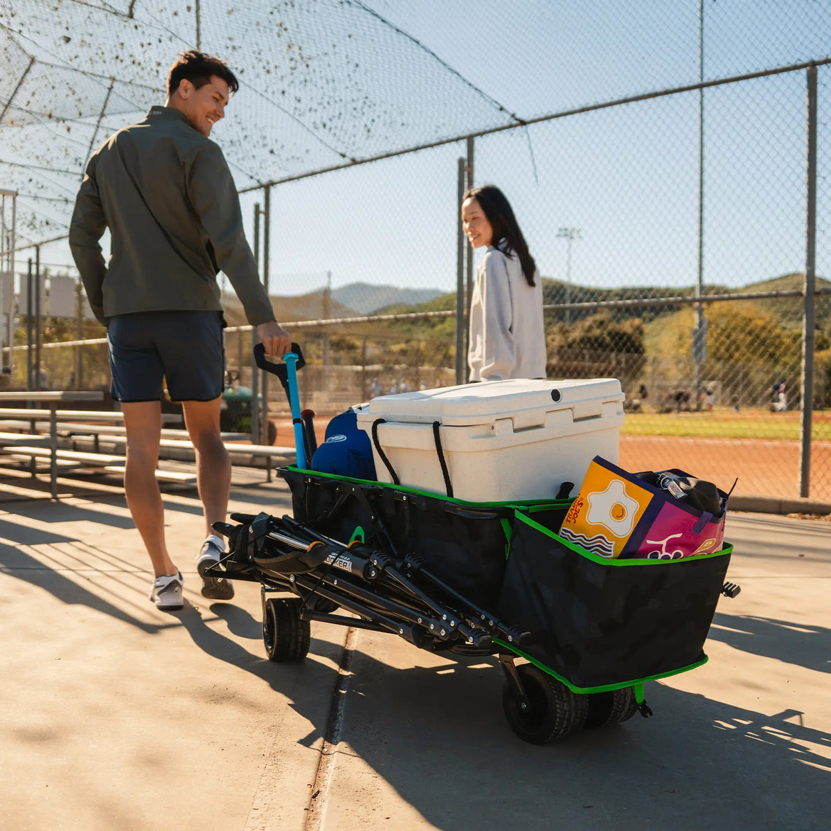 A couple walking together near a baseball field as they pull along a loaded Go-Hauler. 