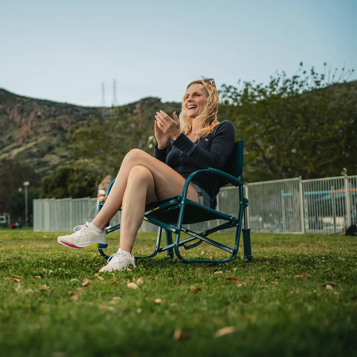 A woman clapping and sitting in a teal Grab & Go Rocker in a grassy area. 