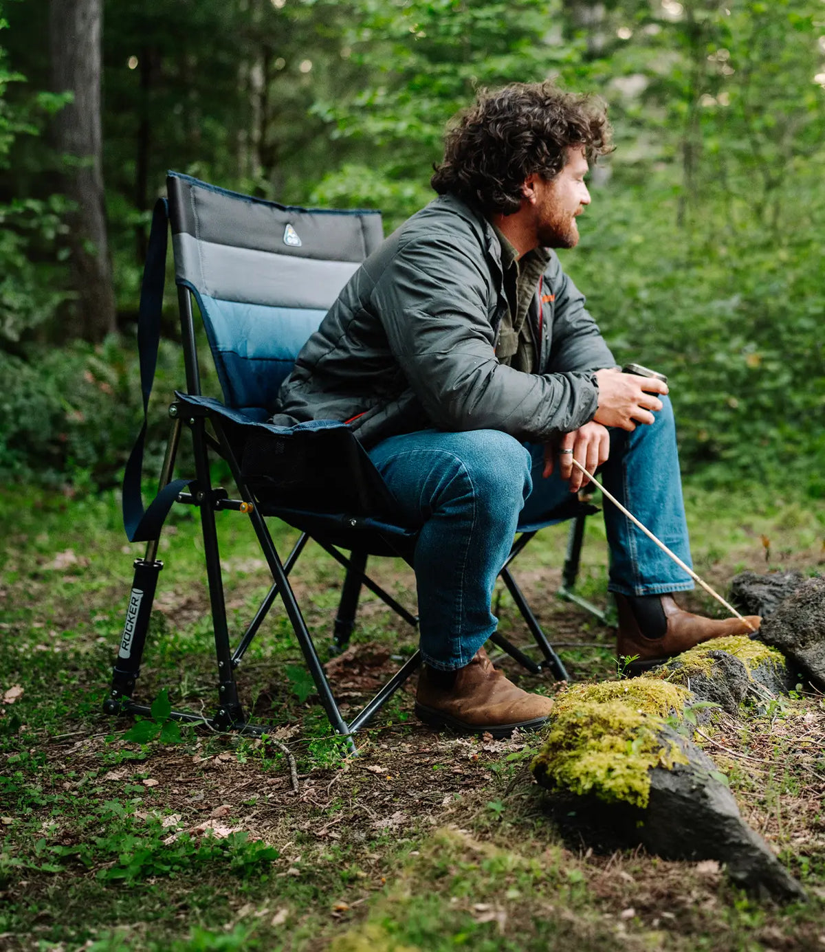 A man sits in a blue and gray Insulated Comfort Pro Rocker in a forest setting, looking off-camera while holding a rope. The chair has a visible Mammut logo on the side panel.