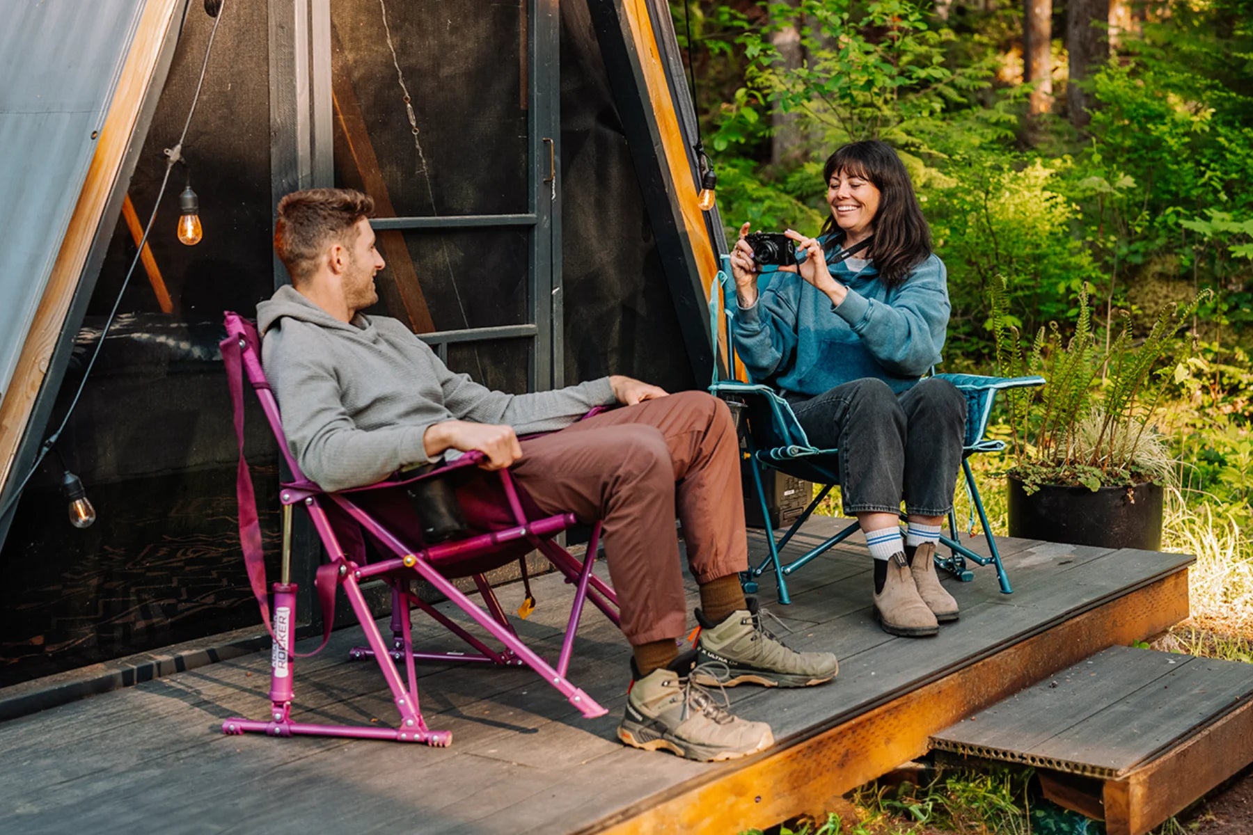 A woman photographs a man relaxing in a berry Insulated Kickback Rocker camp chair, while she sits in a teal version of the same chair on a deck outside a cabin.