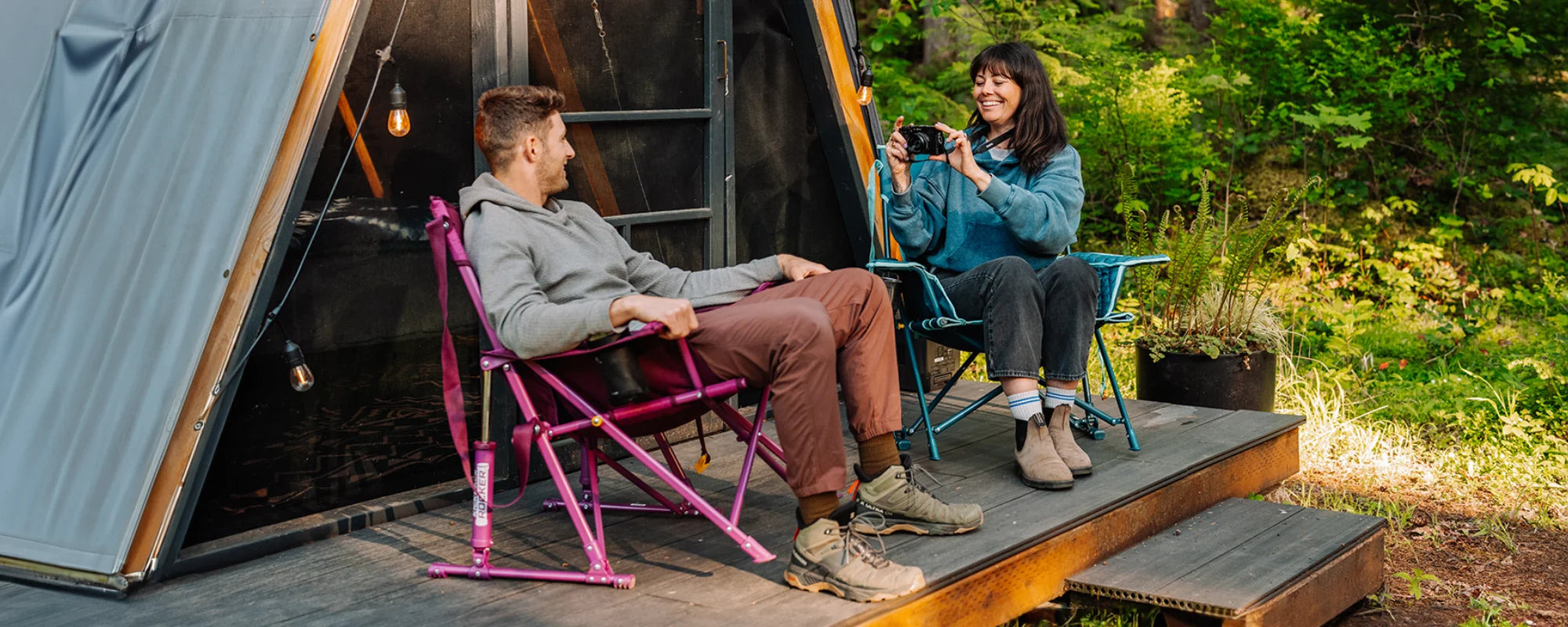 A woman photographs a man relaxing in a berry Insulated Kickback Rocker camp chair, while she sits in a teal version of the same chair on a deck outside a cabin.