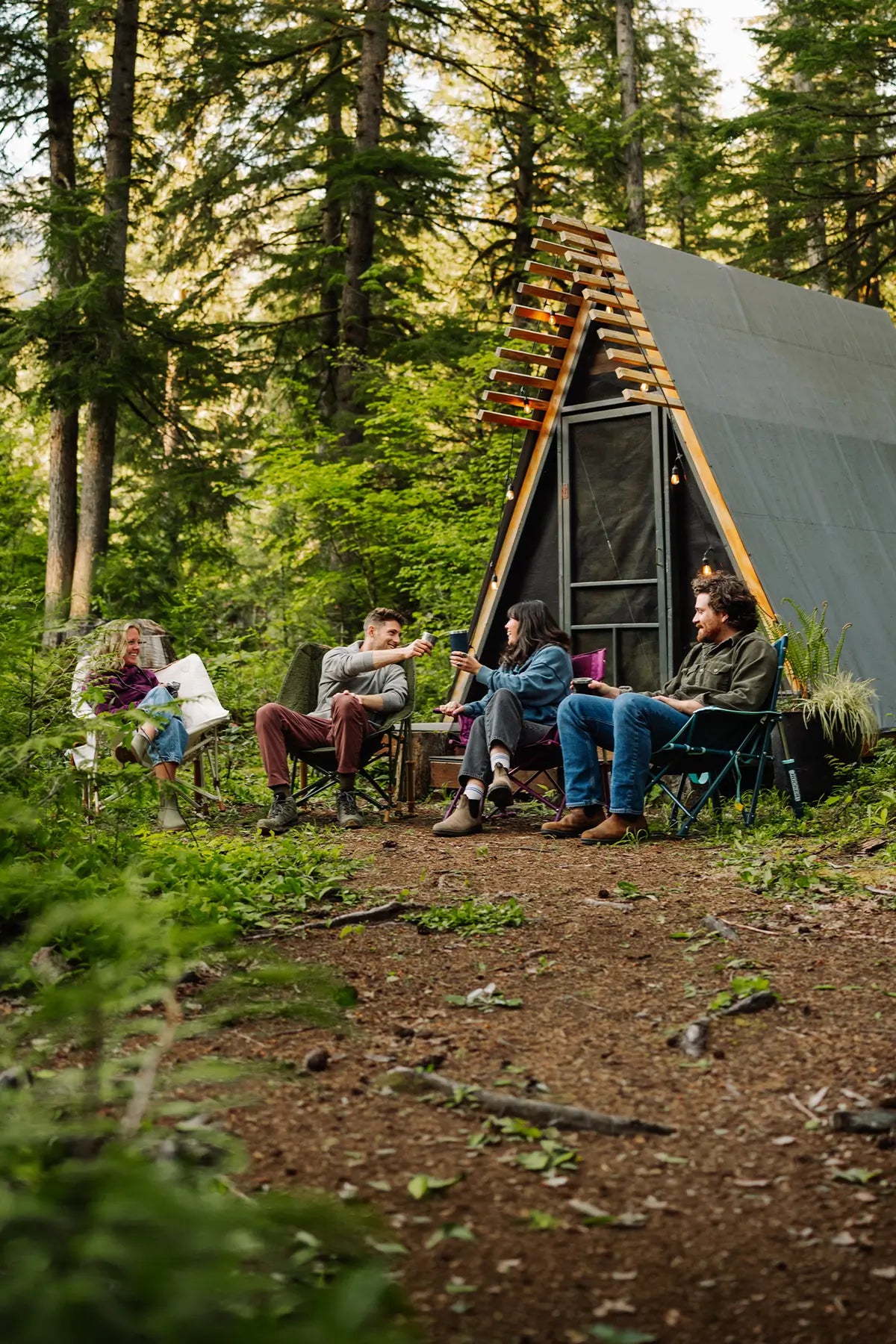 A group of four people sitting and relaxing outdoors in a forest setting next to a modern A-frame cabin with a dark gray roof and glass front door.