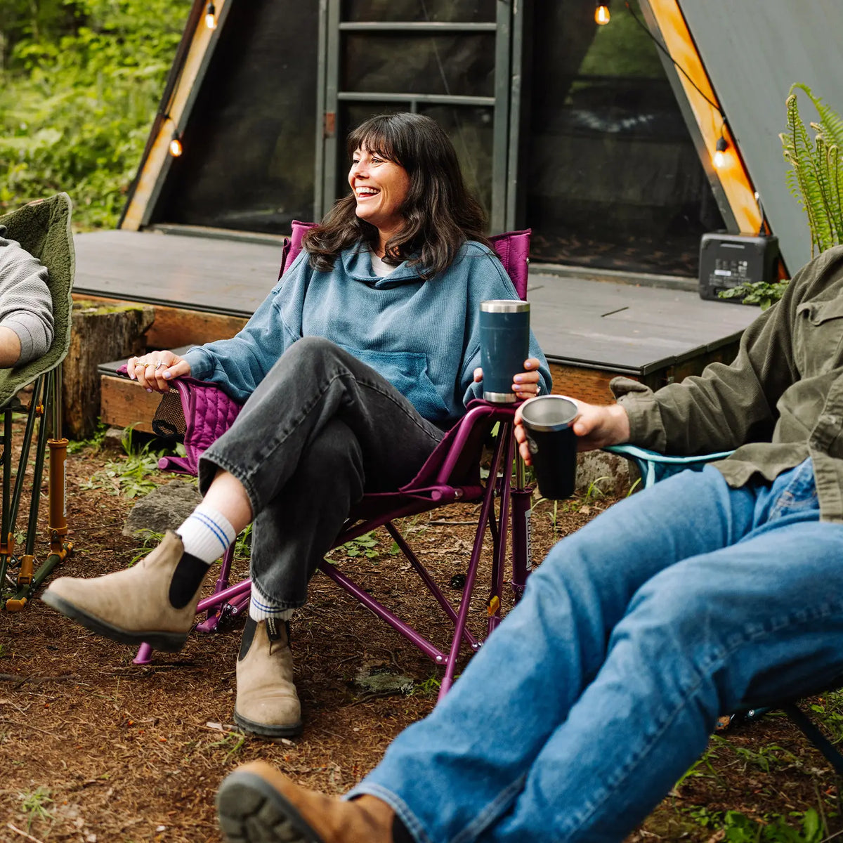 A woman sits in a purple Insulated Kickback Rocker chair in front of an A-frame tent, holding a blue travel tumbler. She is wearing a blue hoodie, dark jeans, and tan boots.