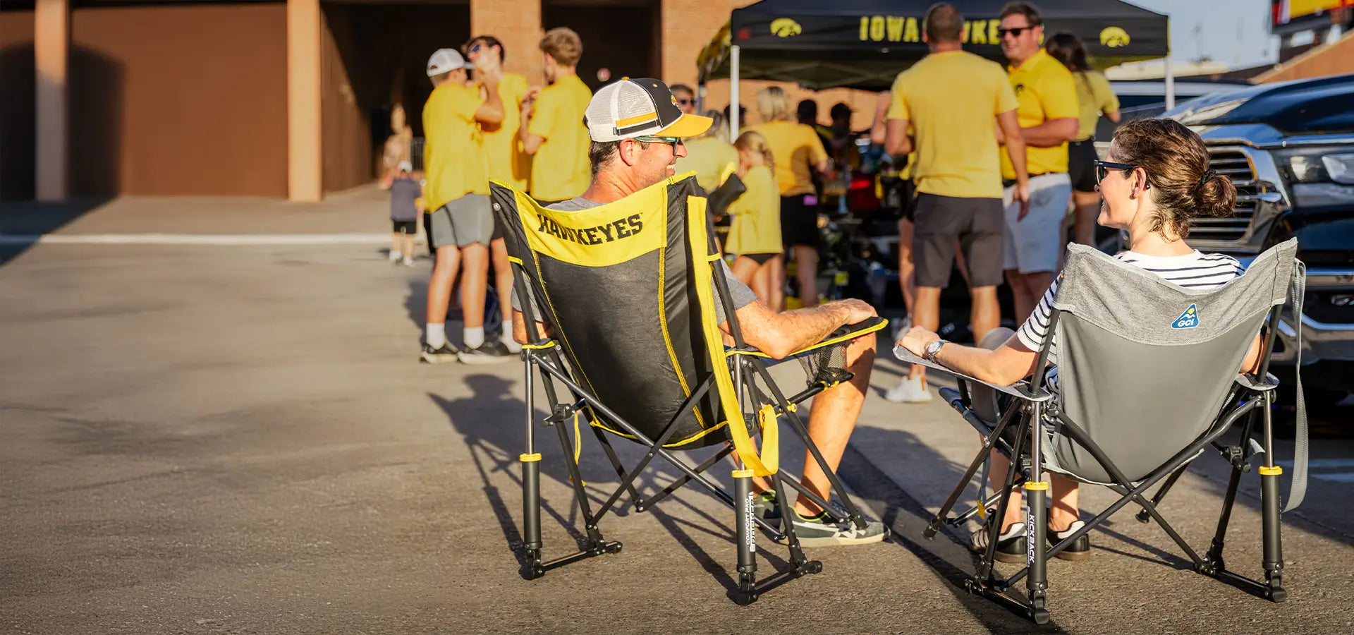 A couple sitting in iowa state comfort pro rocker chairs, that say hawkeyes on the back while hanging out at a tailgate. 