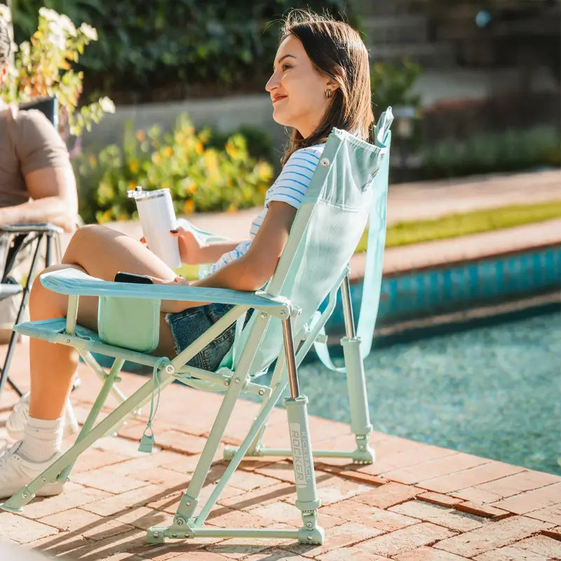 Woman lounging in a pastel green Kickback Rocker by the pool, holding a tumbler and enjoying the sun in a relaxed outdoor setting.